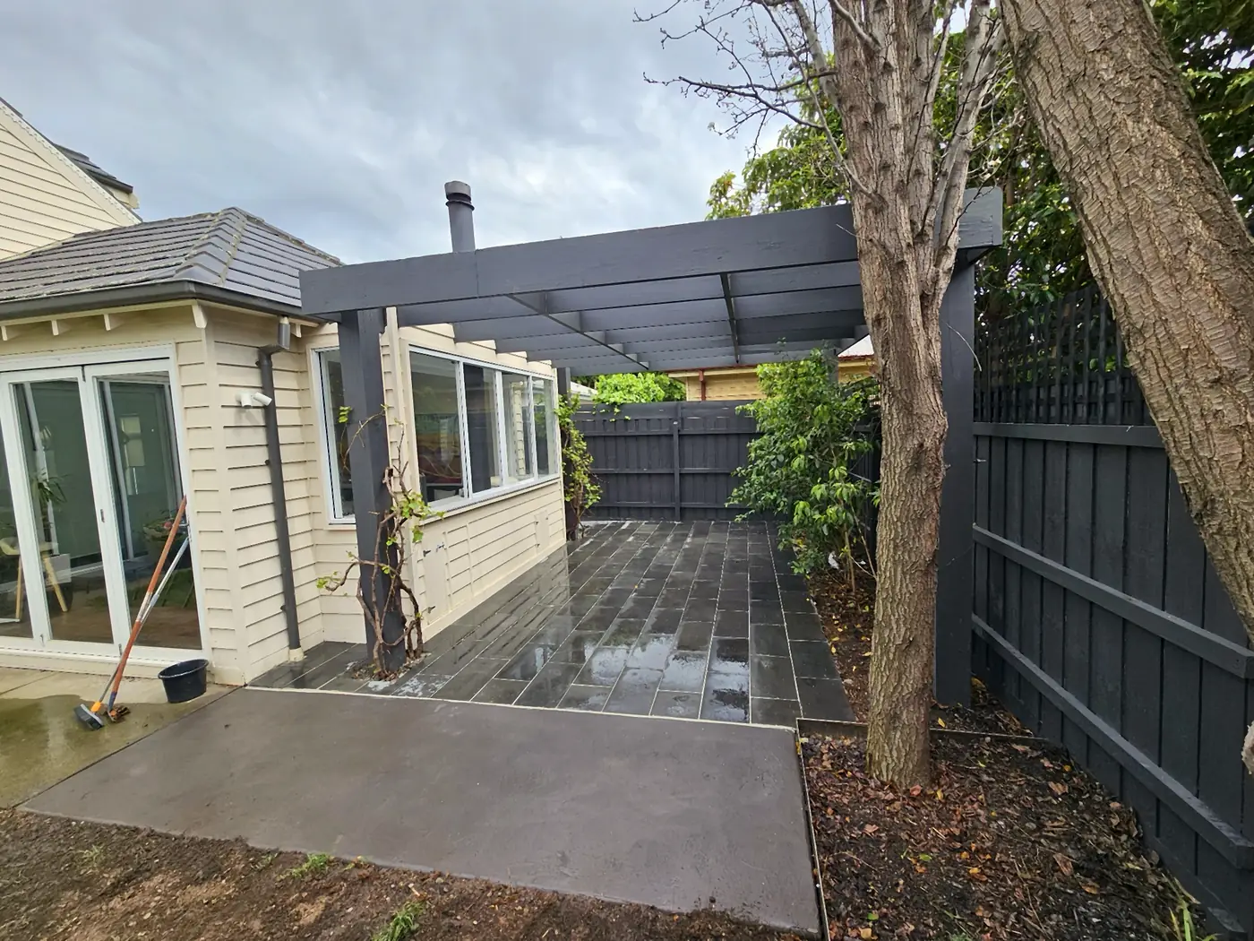 Courtyard with grey paving, a pergola and dark fencing beside a weatherboard home