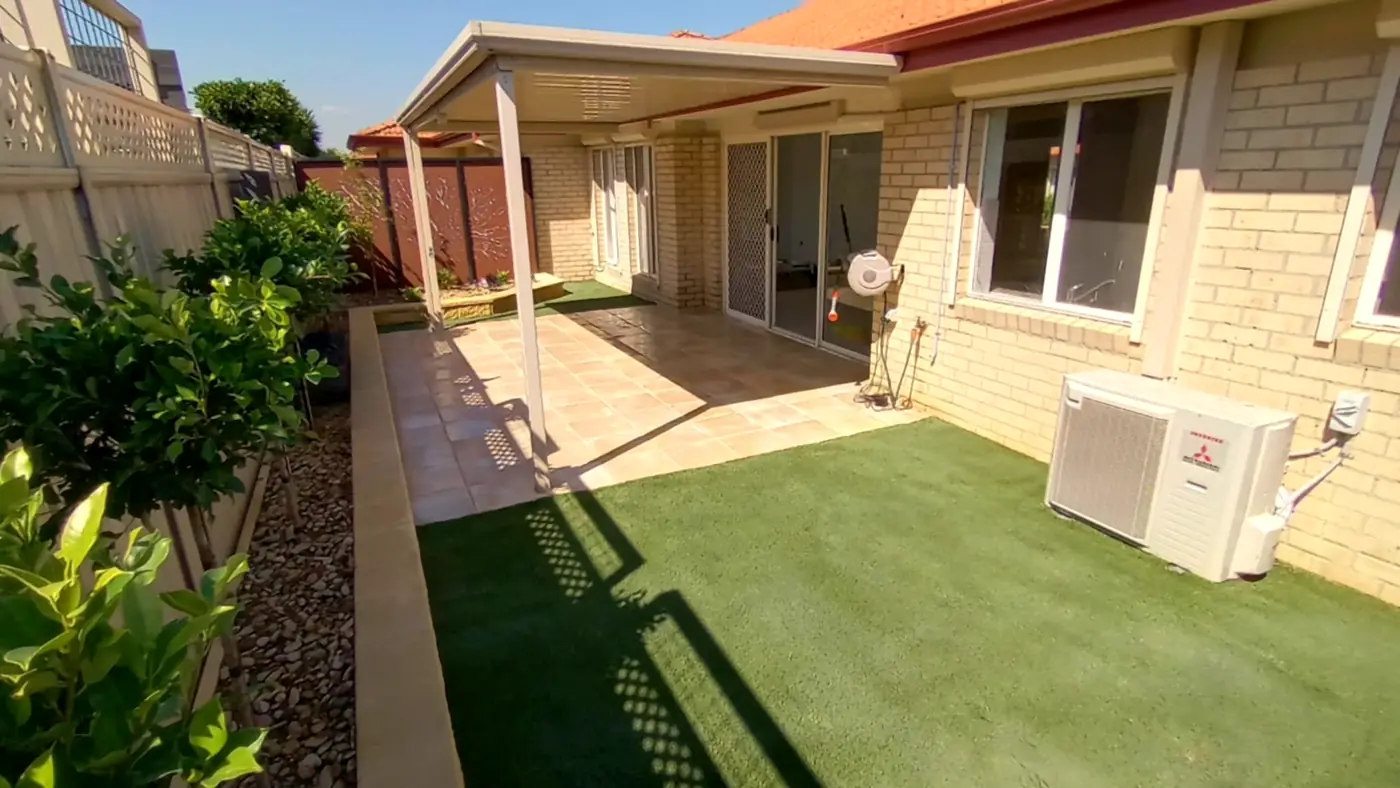 Synthetic turf beside a paved patio and planted garden beds behind a brick home